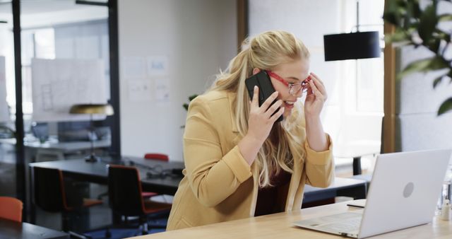 Blonde businesswoman talking on cell phone while working on laptop in modern office. Indicates work stress, multitasking, busy work environment, and high productivity demands. Suitable for articles on workplace stress, career challenges, business communication, professional life, and office settings.