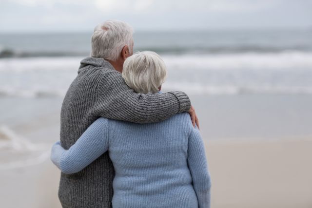 Senior couple standing together on the beach, looking at the ocean while embracing. They are wearing sweaters and have grey hair, evoking a sense of warmth and togetherness in their retirement years. Ideal for use in advertisements or articles about retirement, love in later life, travel, health and wellbeing for seniors, and peaceful lifestyles.