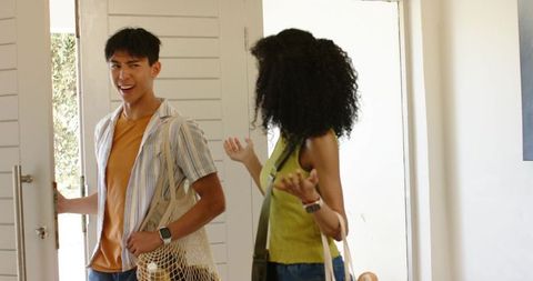 Diverse couple entering modern foyer carrying mesh tote with baguette and canvas tote