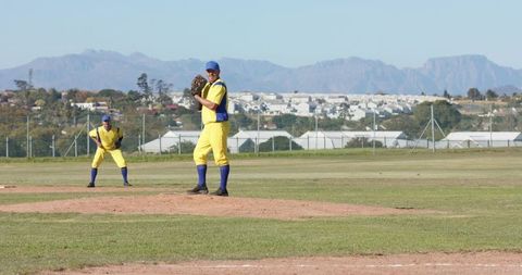 Pitcher and Infielder Wearing Yellow Uniforms Ready on Baseball Mound with Urban Backdrop
