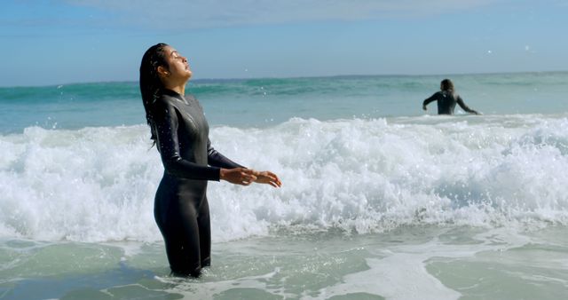 Two women are in the ocean, wearing wetsuits and enjoying the waves. One woman appears to be taking in the refreshing sea air, while the other is in the background partially in the water. Perfect for themes related to beach vacations, water sports, outdoor activities, and refreshing moments in nature.