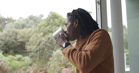 African American man enjoying morning coffee by sliding door overlooking garden in rust sweater