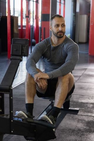 Mid-adult indian man sitting on rowing machine resting between sets in gym workout