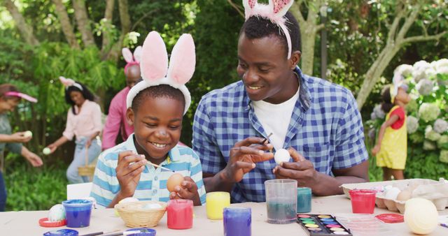 Father and son are sitting at an outdoor table, painting Easter eggs while wearing bunny ears. The setting is a lush garden, indicating a springtime holiday celebration. This cheerful and vibrant moment showcases family bonding, creativity, and festive activities that can be perfect for illustrating family-oriented, holiday celebration articles, advertisements for Easter products, or promoting artistic activities.