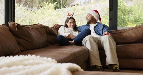 Diverse couple relaxing on sectional sofa wearing santa hat and reindeer headband