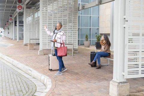 Senior african american woman waving for ride with luggage at curbside transit shelter