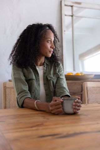 African american woman sitting at wooden dining table holding ceramic mug looking contemplative