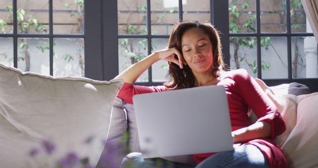 A woman is sitting comfortably on a couch at home while using a laptop. She is smiling and relaxed, creating a positive and cozy atmosphere. This content is great for articles or blogs about remote work, home offices, relaxation, work-life balance, and modern lifestyle. Companies can also use it for promotions related to tech gadgets, casual wear, or home decor.