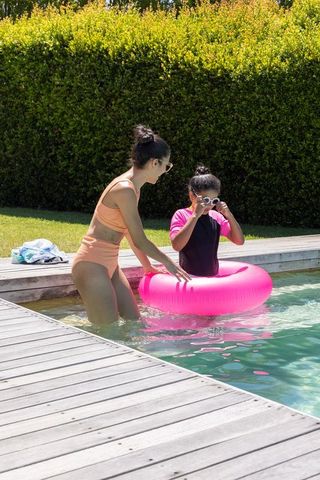 African american woman helping child adjust swim goggles with pink inflatable ring at pool