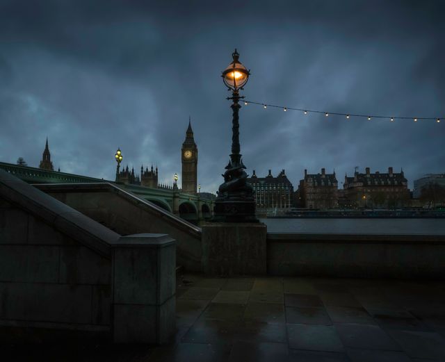 Night view captures iconic Big Ben and Westminster Bridge under cloudy sky with street lamp lit in foreground. London's famous landmarks illuminated against dark landscape, suitable for travel blogs, tourism promotions, cityscape wall art, and historic architecture features.