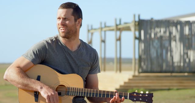 Man sitting outside on a sunny day playing guitar. This casual scene is perfect for themes related to music, relaxation, and outdoor activities. Can be used for advertisements, blog posts, and social media promotions focused on lifestyle, leisure, and creativity.