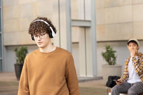 Curly-haired young man walking with headphones on urban campus while friend using phone