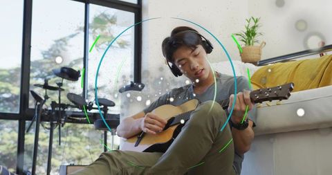 Korean teen playing acoustic guitar in sunlit living room home studio with headphones