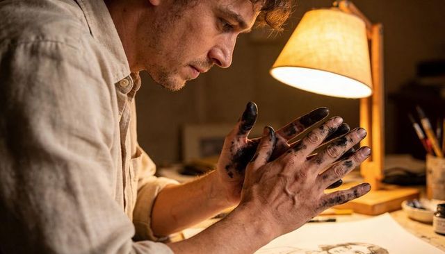 Male artist leaning with ink-stained hands focusing on portrait sketch by warm lamp