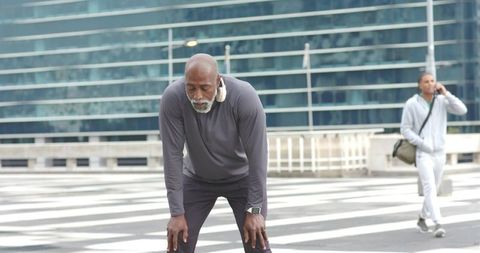 Senior man leaning on knees at urban crosswalk after outdoor workout wearing headphones