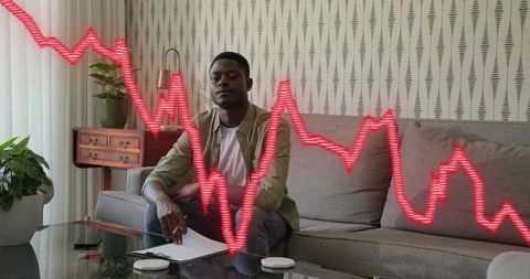 Man sitting on sofa writing in notebook with red falling stock chart overlay, remote work
