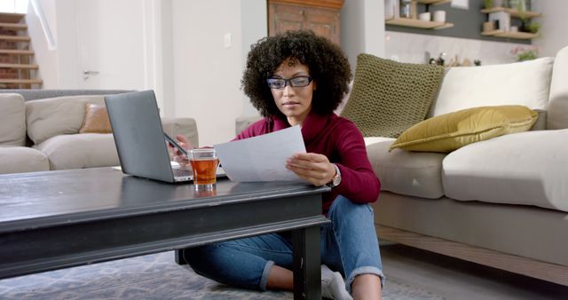 Young woman sitting on floor in living room, working on laptop and holding documents. Ideal for content related to remote work, home office setups, work-life balance, and productivity tips.