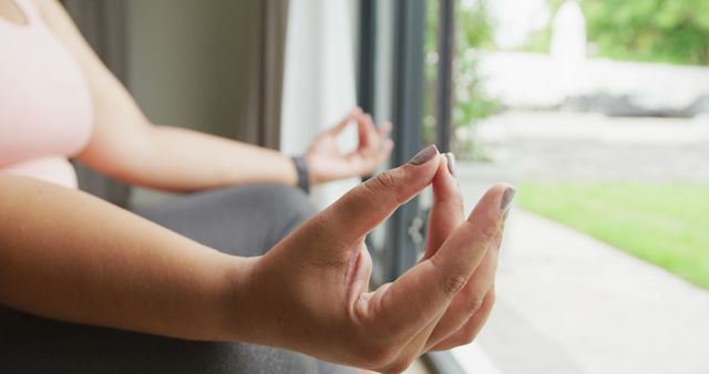 Close-up of person sitting with hands in lotus position, practicing meditation near open window with garden view. Ideal for articles or blogs on mindfulness, mental health, yoga practice, and holistic living.