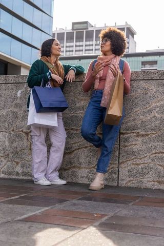 Two friends chatting and holding shopping bags while leaning on stone ledge in city plaza