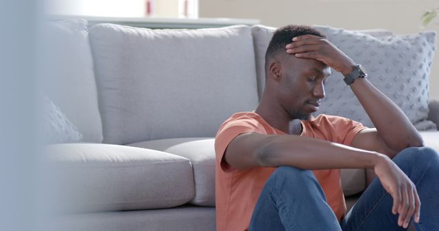 Young man is sitting on the floor with hand on head, appearing stressed and thoughtful. The setting is a living room with a sofa in the background. Useful for concepts such as mental health, emotional struggle, stress, and solitude.