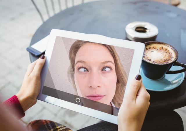 Woman making humorous faces during a video call on digital tablet at outdoor cafe. Tablet reflects close-up of her funny expressions. Coffee cup on table adds to relaxed ambiance. Ideal for uses in articles regarding video communication, technology in daily life, or casual moments of joy.