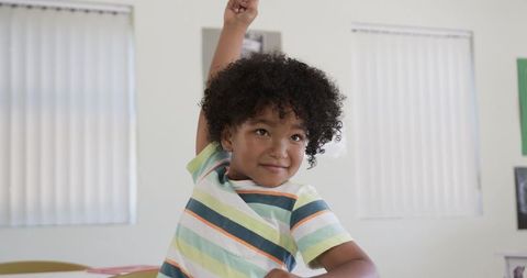 Smiling student raising right hand at classroom desk wearing pastel-striped tee