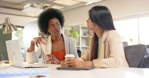 Female Colleagues Collaborating at Modern Office Desk Discussing Ideas Over Coffee