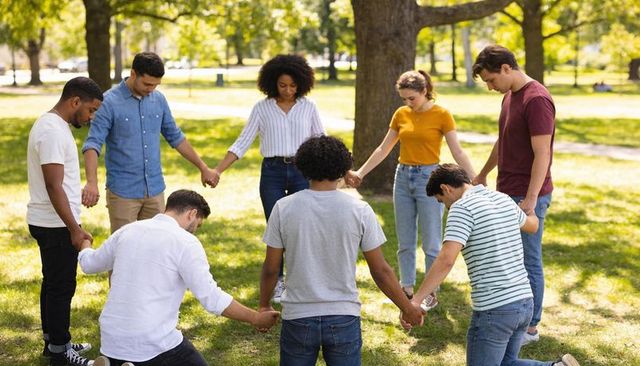 Diverse group holding hands and bowing heads in sunlit park circle
