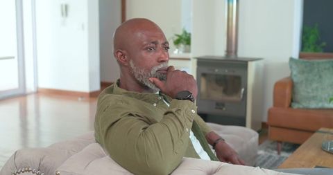 Middle-aged man sitting on tufted sofa wearing olive shirt resting chin in cozy living room