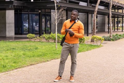 African american commuter wearing orange sweater listening to headphones on campus walkway