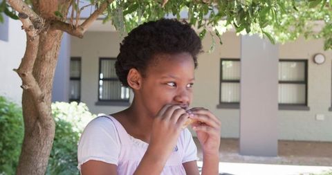 African girl sitting under tree eating sandwich in courtyard enjoying outdoor snack