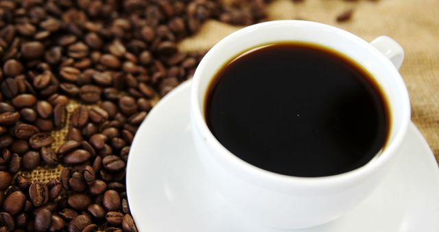 A cup of black coffee sits on a saucer surrounded by roasted coffee beans, with copy space. Its rich color and the scattered beans suggest a fresh, aromatic brew perfect for coffee enthusiasts.