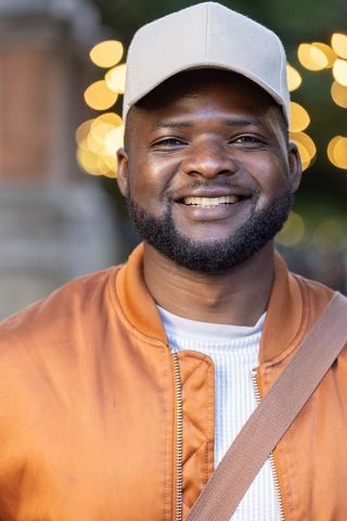 African man smiling at dusk wearing beige cap and orange jacket with urban bokeh lights