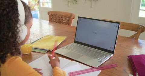 Young student doing distance learning at home wearing headphones and writing by laptop