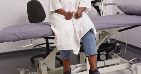 African american woman sitting on medical exam table wearing gown clasping hands clinic