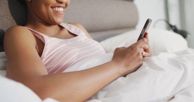 Happy african american woman laying in bed, using smartphone in bedroom. domestic lifestyle, spending free time at home.
