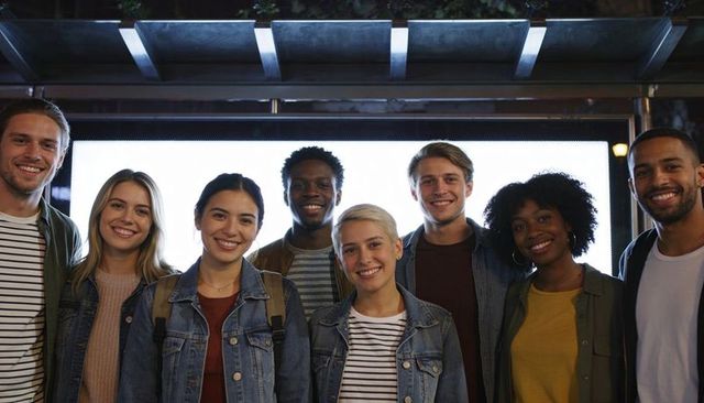 Diverse young friends smiling under backlit transit shelter at night, casual denim