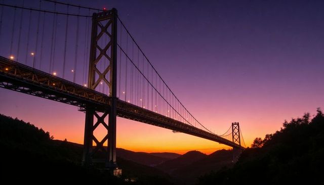 Twilight suspension bridge spanning valley, lit roadway glowing against purple orange sky