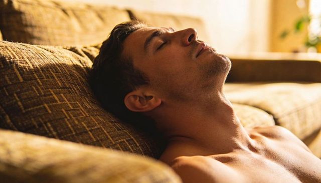 Shirtless man reclining on sunlit sofa, resting in warm golden-hour living room light