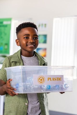 African american boy holding plastic recycling bin with bottles in classroom