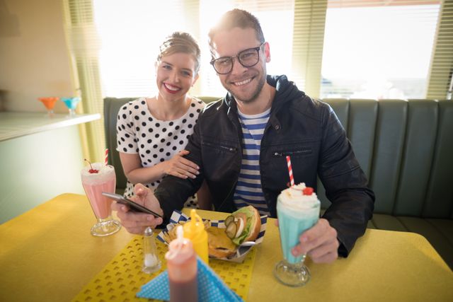 Young couple sitting in a retro diner, enjoying milkshakes and using a mobile phone. They are smiling and appear to be having a good time. This image can be used for advertisements, social media posts, or articles related to dating, lifestyle, food, and vintage themes.