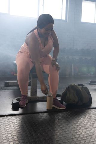 African american woman reaching for water bottle on bench during gym workout, training