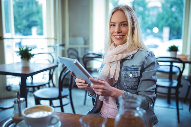 Young woman sitting in cozy cafe, using tablet while smiling at camera. Ideal for content related to technology, social media, leisure activities, cafe culture, modern lifestyle, and digital communication.