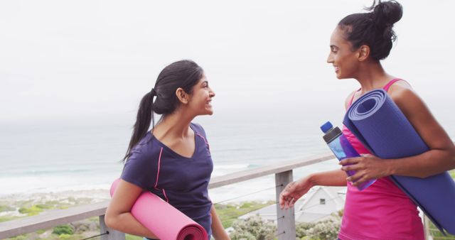 Two women wearing sportswear holding yoga mats and bottle, having cheerful conversation on wooden terrace with ocean view in background. Ideal for promoting active lifestyle, fitness routines, outdoor activities and female friendships.