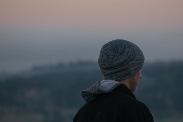 A young man wearing a gray beanie and hoodie gazing at the sunset. Ideal for themes relating to contemplation, solitude, and serenity. Suitable for blogs, motivational content, mental health articles, and nature appreciation media.