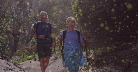 Sunlit senior couple walking forest trail with backpacks and trekking poles, active retirement