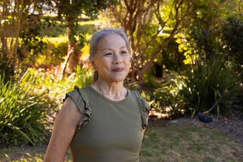 Senior asian woman standing in sunlit backyard garden wearing olive green ruffled top