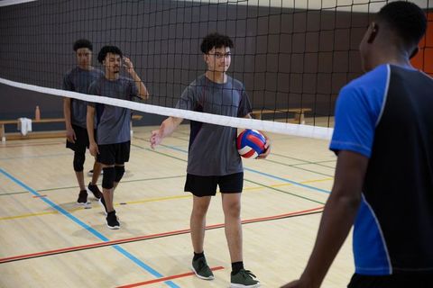 High school diverse volleyball team shaking hands over net while holding ball in indoor gym