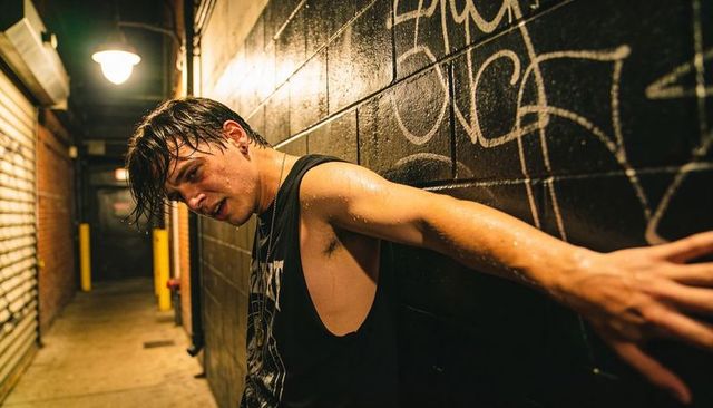 Leaning young man sweating in tank top pressing arm on graffiti-covered wall in moody urban alley