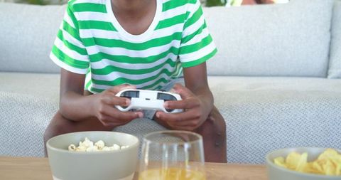 African american boy playing video games on couch with snacks and drink